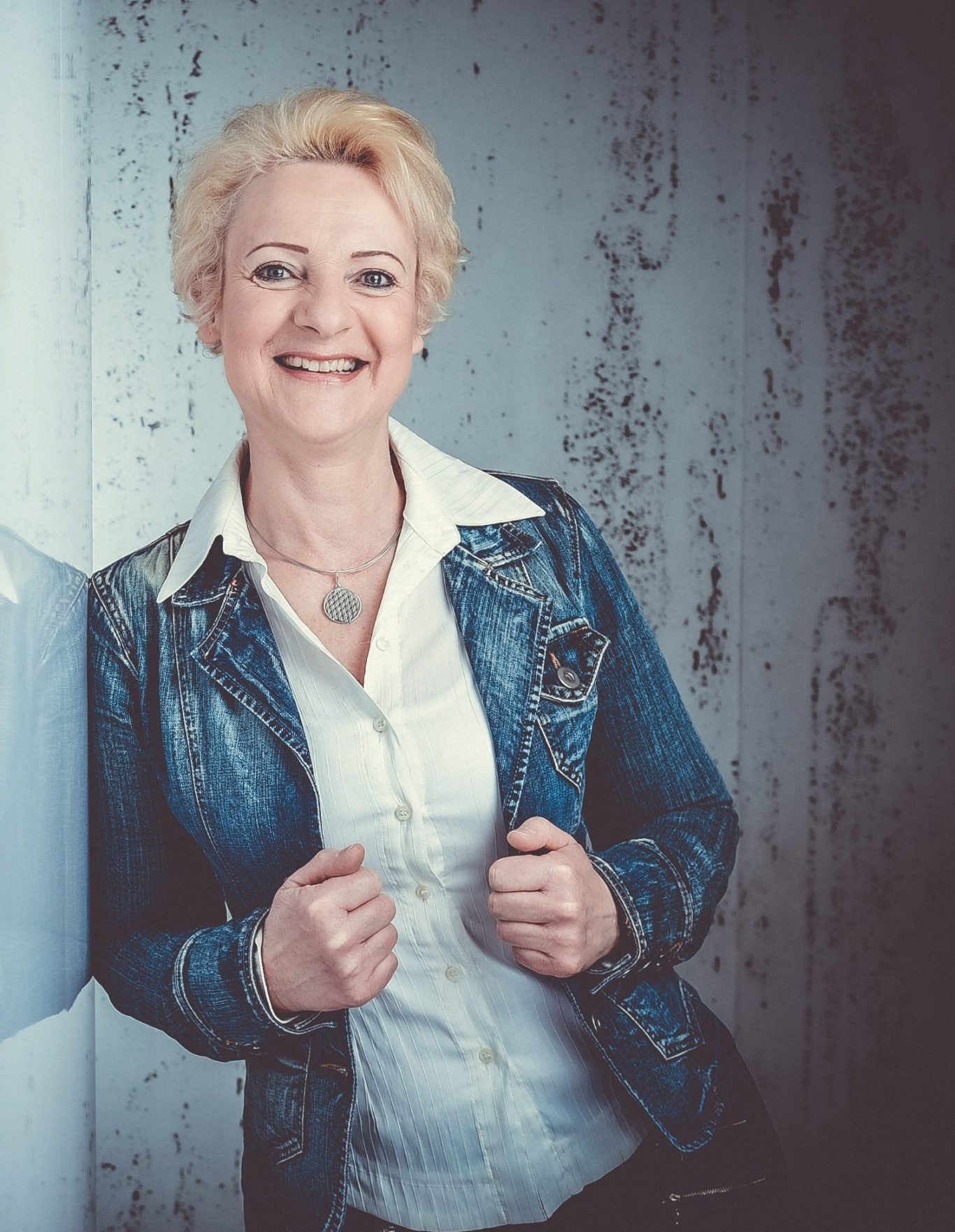 Astrid Steffen Eine Frau gekleidet in einer weißen Bluse, darüber eine Jeansjacke. Sie lehnt sich an einer Wand an und lächelt freunlich in die Kamera.
